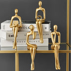 Four abstract golden figures are arranged, sitting on a stack of books and a gold-framed glass shelf.