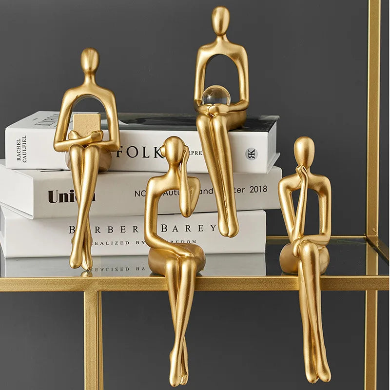 Four abstract golden figures are arranged, sitting on a stack of books and a gold-framed glass shelf.