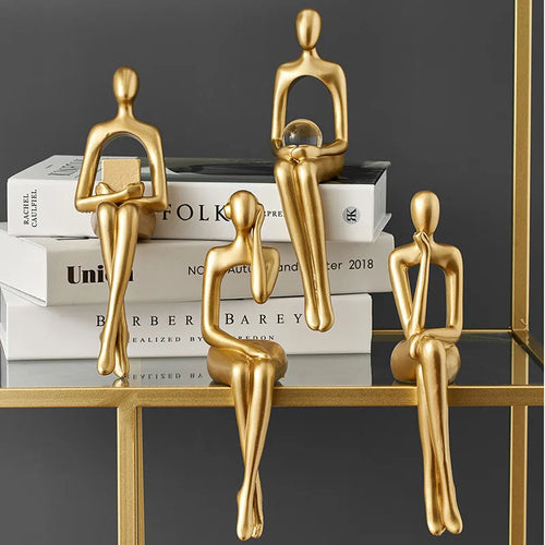 Four abstract golden figures are arranged, sitting on a stack of books and a gold-framed glass shelf.