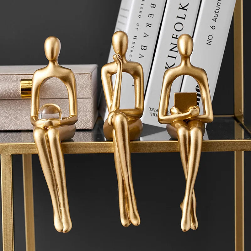 Three abstract gold figurines sit on a glass shelf, one holding a clear orb, another thinking, and the third holding a book.