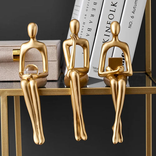 Three abstract gold figurines sit on a glass shelf, one holding a clear orb, another thinking, and the third holding a book.