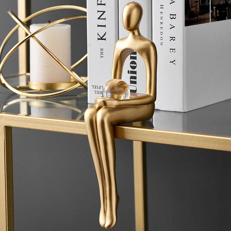 Gold abstract sitting figurine with a hollow torso, holding a clear sphere while perched on a gold and glass shelf.