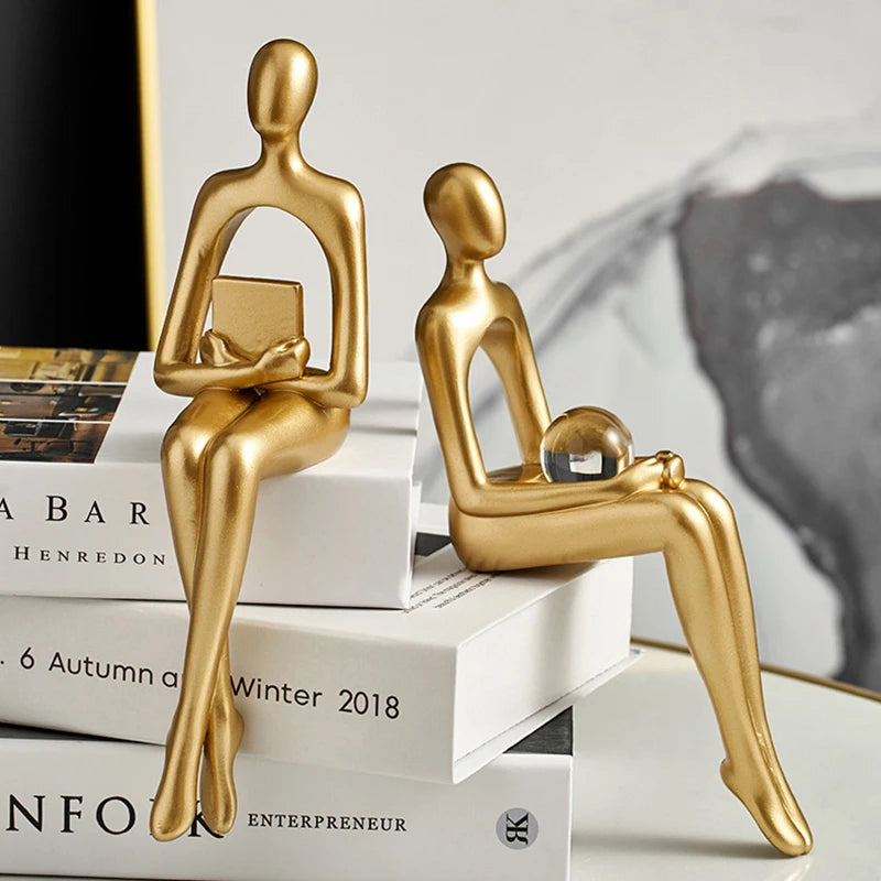Two abstract golden figurines sit on stacked books; one holds a small book, the other a clear sphere.