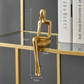 Golden abstract sitting figurine, sculpted in a thoughtful pose, resting on a glass shelf with books in the background.