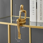Golden abstract sitting figurine, sculpted in a thoughtful pose, resting on a glass shelf with books in the background.