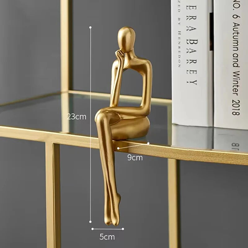 Golden abstract sitting figurine, sculpted in a thoughtful pose, resting on a glass shelf with books in the background.