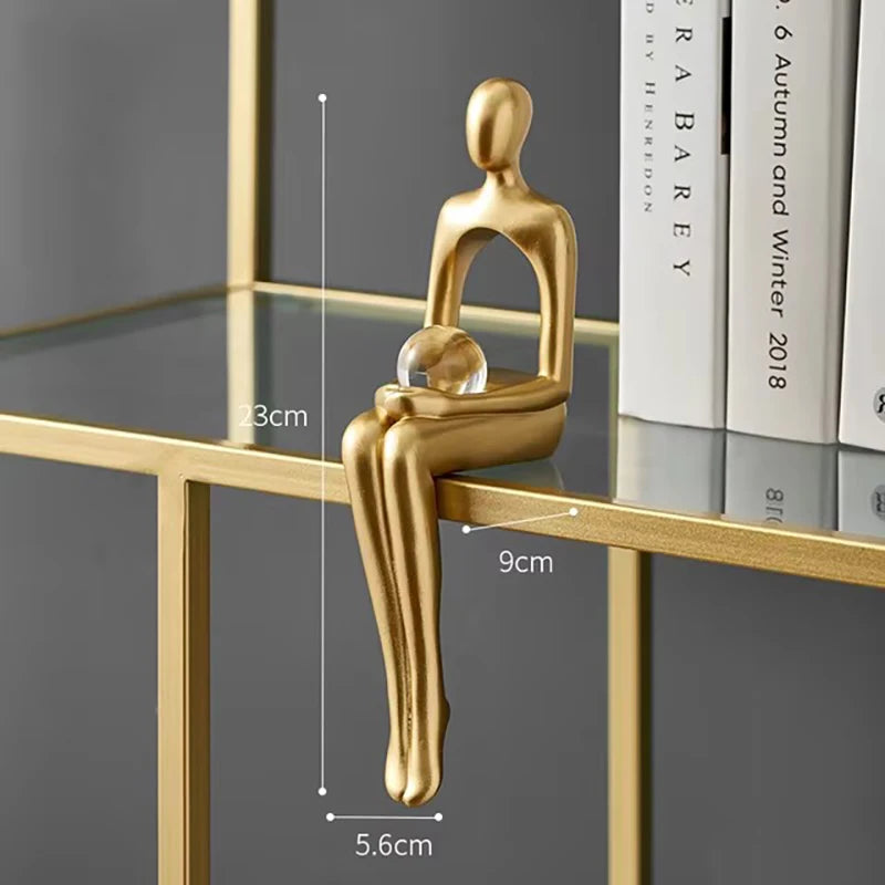 Abstract gold-toned human-shaped figurine sitting on a gold and glass shelf, holding a clear sphere between its hands.