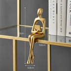 Golden abstract figurine of a person sitting on a shelf edge, holding a small book.