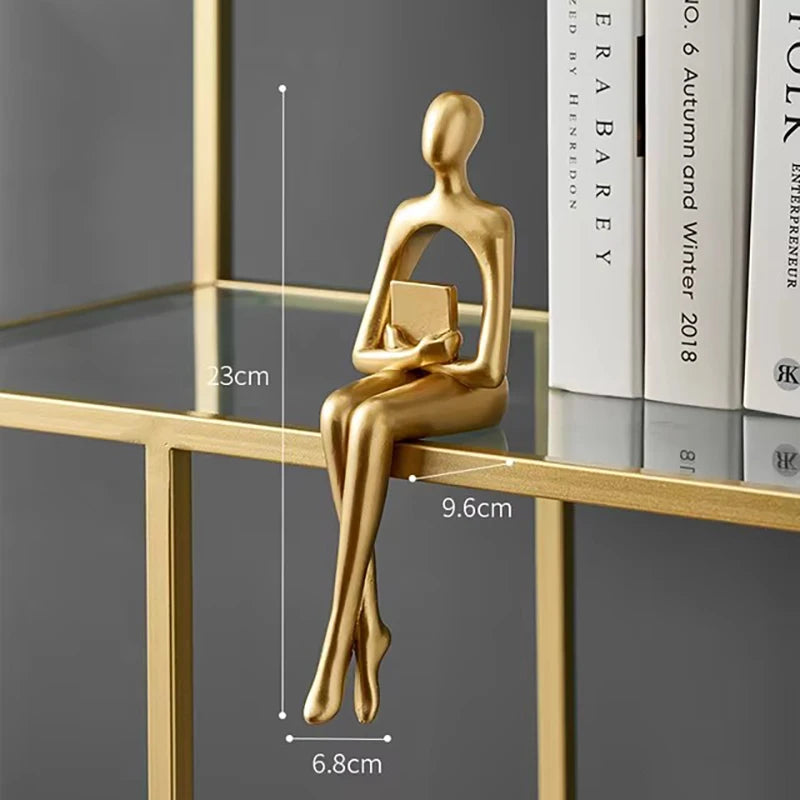 Golden abstract figurine of a person sitting on a shelf edge, holding a small book.