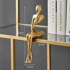 Abstract gold metallic figurine with an arm covering its face, sitting on a glass shelf with books nearby.