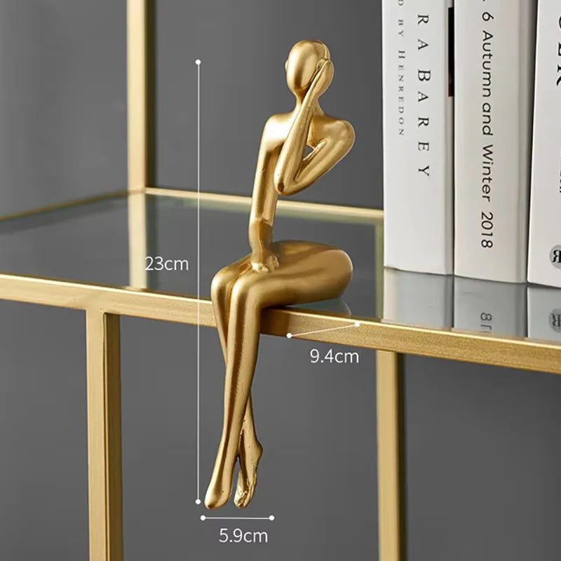 Abstract gold metallic figurine with an arm covering its face, sitting on a glass shelf with books nearby.