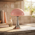 Pink mushroom-shaped glass lamp with a polished silver flared base on a rustic wooden countertop by a sunny window.