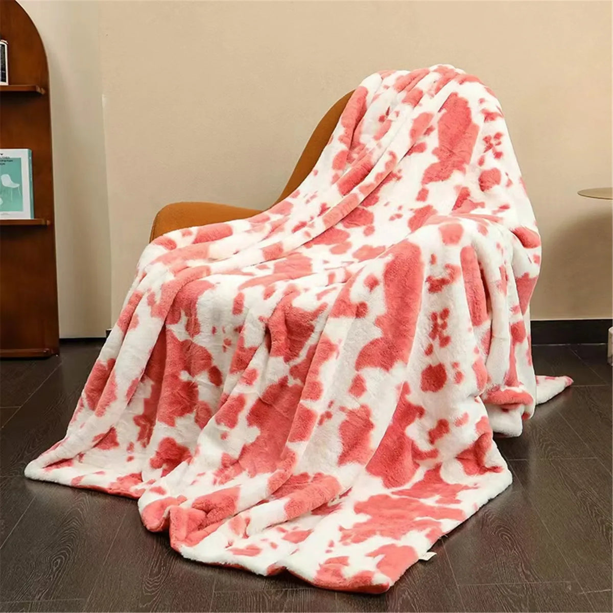A soft, plush white blanket with large pink cow print spots draped over a brown armchair on a dark wood floor.