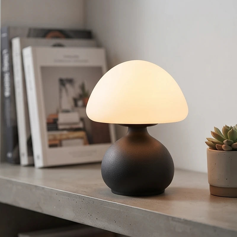 Mushroom-shaped table lamp with a glowing white frosted shade and a dark, textured base on a grey shelf.