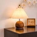 A pleated fabric shade illuminates a round wooden lamp with a chrome accent on a dark dresser, beside a framed bird print.