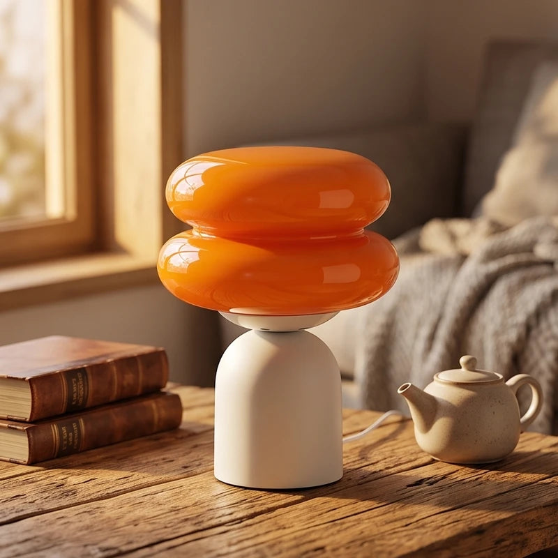 Orange glass lamp with a two-tiered, mushroom-like shade and a rounded off-white base on a wooden table.