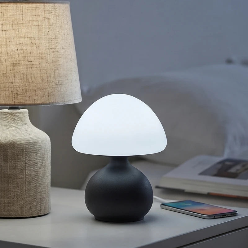 A mushroom-shaped table lamp with a glowing white dome top and a dark gray base sits on a nightstand.