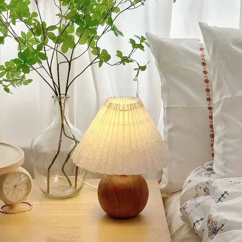 Illuminated table lamp with a pleated conical shade and a round wooden base, beside a vase of green leaves.