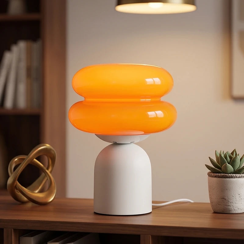 An orange double-tiered glass lamp shade sits atop a white dome base, casting a warm glow on a wooden desk.