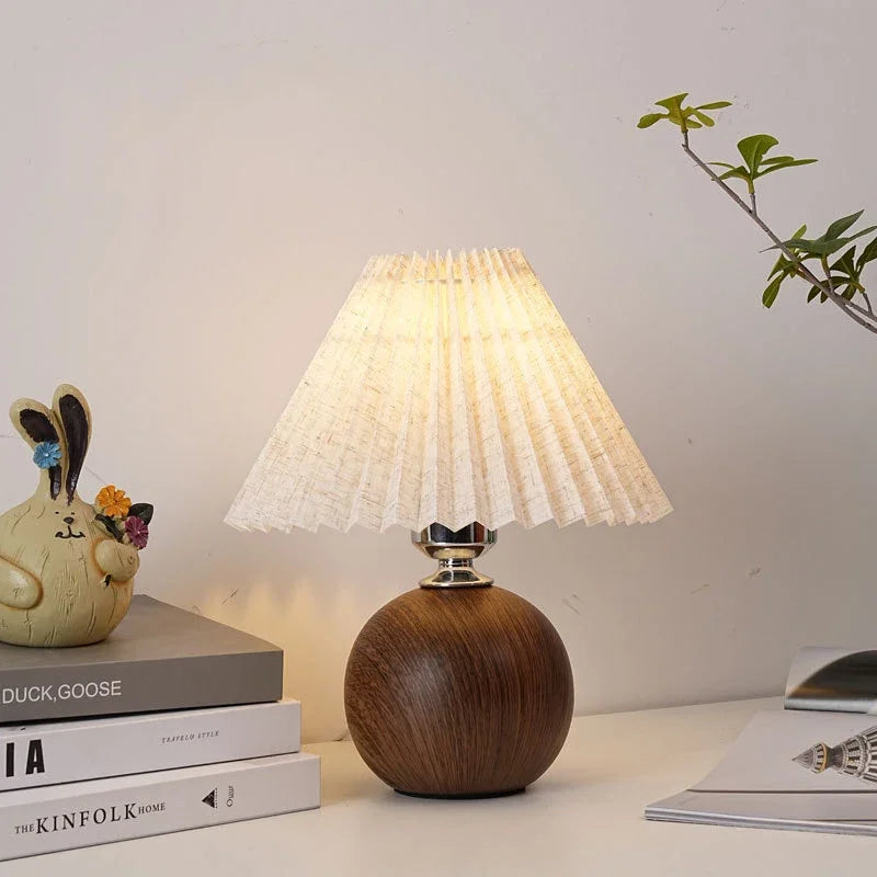 Table lamp with a pleated cream linen shade, a polished silver neck, and a round dark wood grain base.