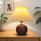 Illuminated table lamp with a dark wood spherical base and a pleated, light yellow fabric shade on a wooden table.