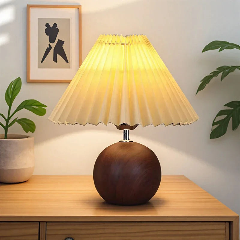 Illuminated table lamp with a dark wood spherical base and a pleated, light yellow fabric shade on a wooden table.