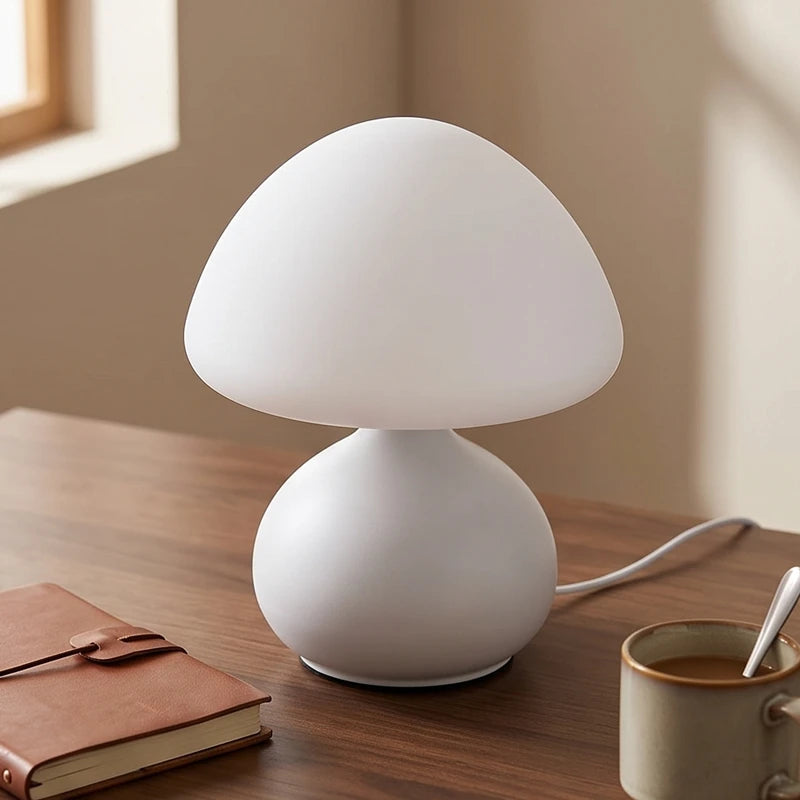 White mushroom-shaped table lamp with a frosted glass shade and base on a wooden desk next to a notebook and coffee mug.
