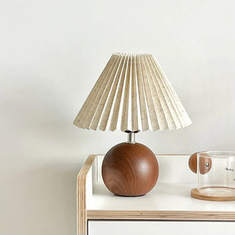 Table lamp with a light beige pleated fabric shade and a round, dark brown wooden base on a white surface.