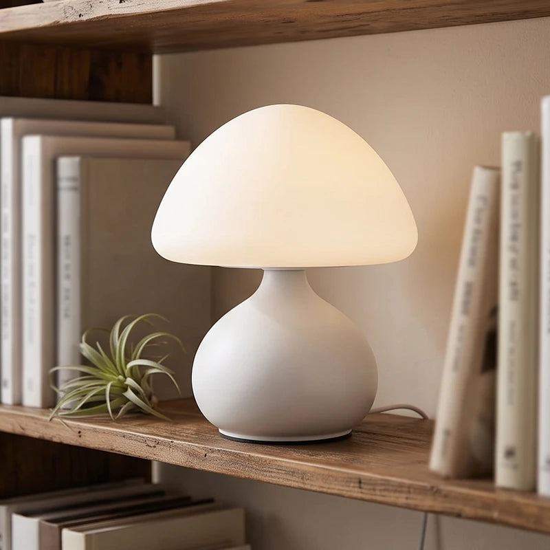 Glowing white mushroom-shaped table lamp with a frosted glass dome and a matte white base, sitting on a rustic wooden shelf surrounded by books and an air plant.