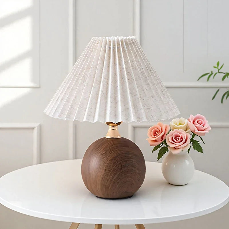 A table lamp with a pleated off-white fabric shade, a round dark wood base, and a white vase with colorful roses.