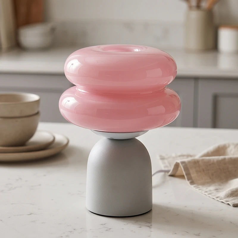 Pink double-bubble glass lamp on a matte light gray arched base, sitting on a white counter with dishes and a cloth.