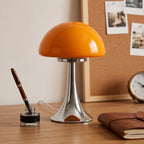 Orange mushroom-shaped lamp with a glossy chrome base on a wooden desk, next to an inkwell and leather notebook.