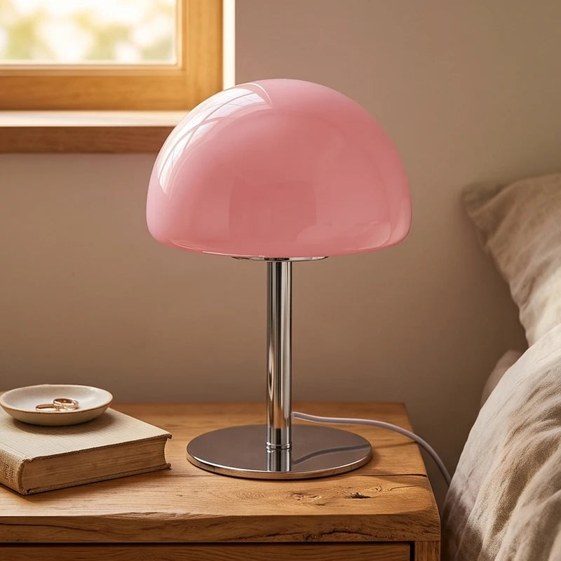 Pink mushroom-shaped glass lamp with a chrome base on a wooden nightstand beside a book and ring dish.