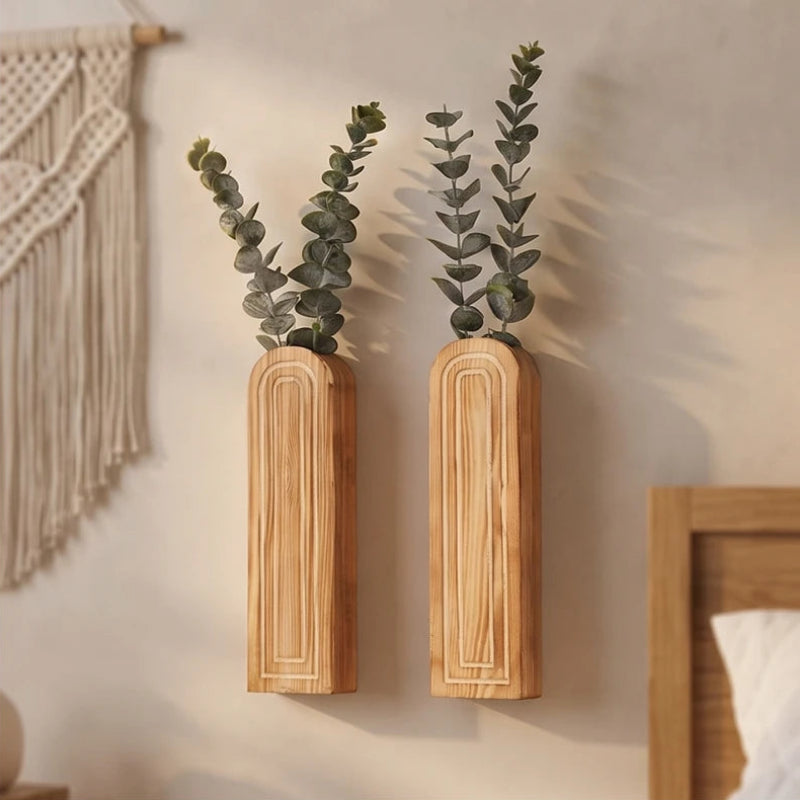 Two light wood wall-mounted vases with carved arch details, holding green leafy sprigs against a neutral wall.