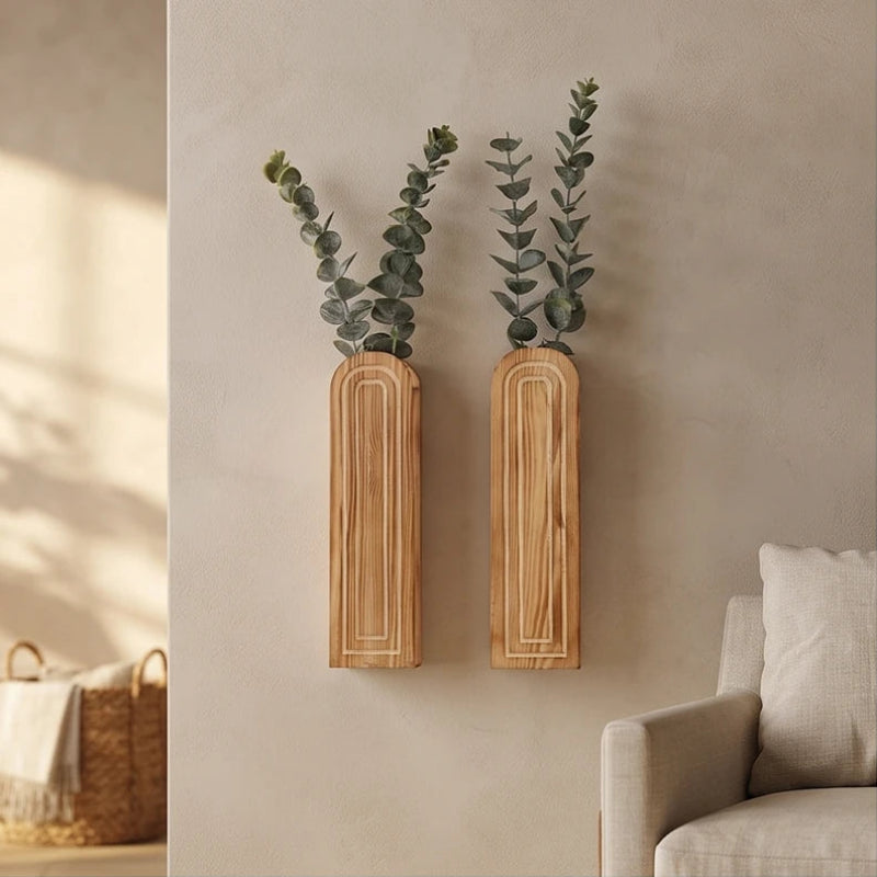Two light wooden rectangular wall vases with carved arch patterns hold leafy green stems on a beige wall.