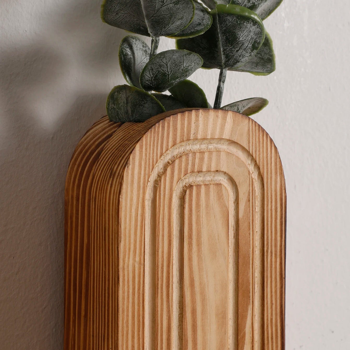 Close-up of a light brown wooden vase with carved arch patterns, holding green eucalyptus leaves against a white wall.