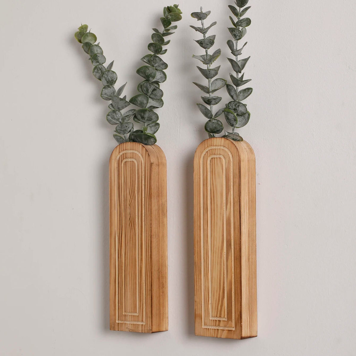 Two light wooden rectangular wall vases with rounded tops, each holding green artificial eucalyptus branches against a white wall.